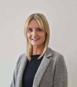 A headshot of Samantha Mitchell smiling at the camera, wearing a black top and grey jacket.